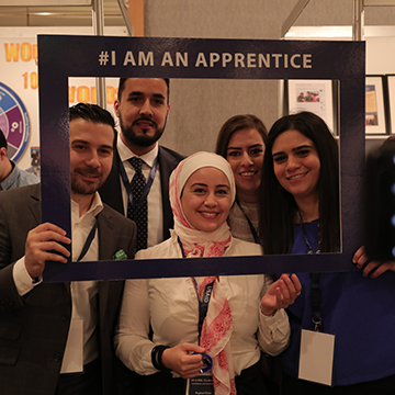 Group of men and women pose behind frame that reads "I Am An Apprentice"